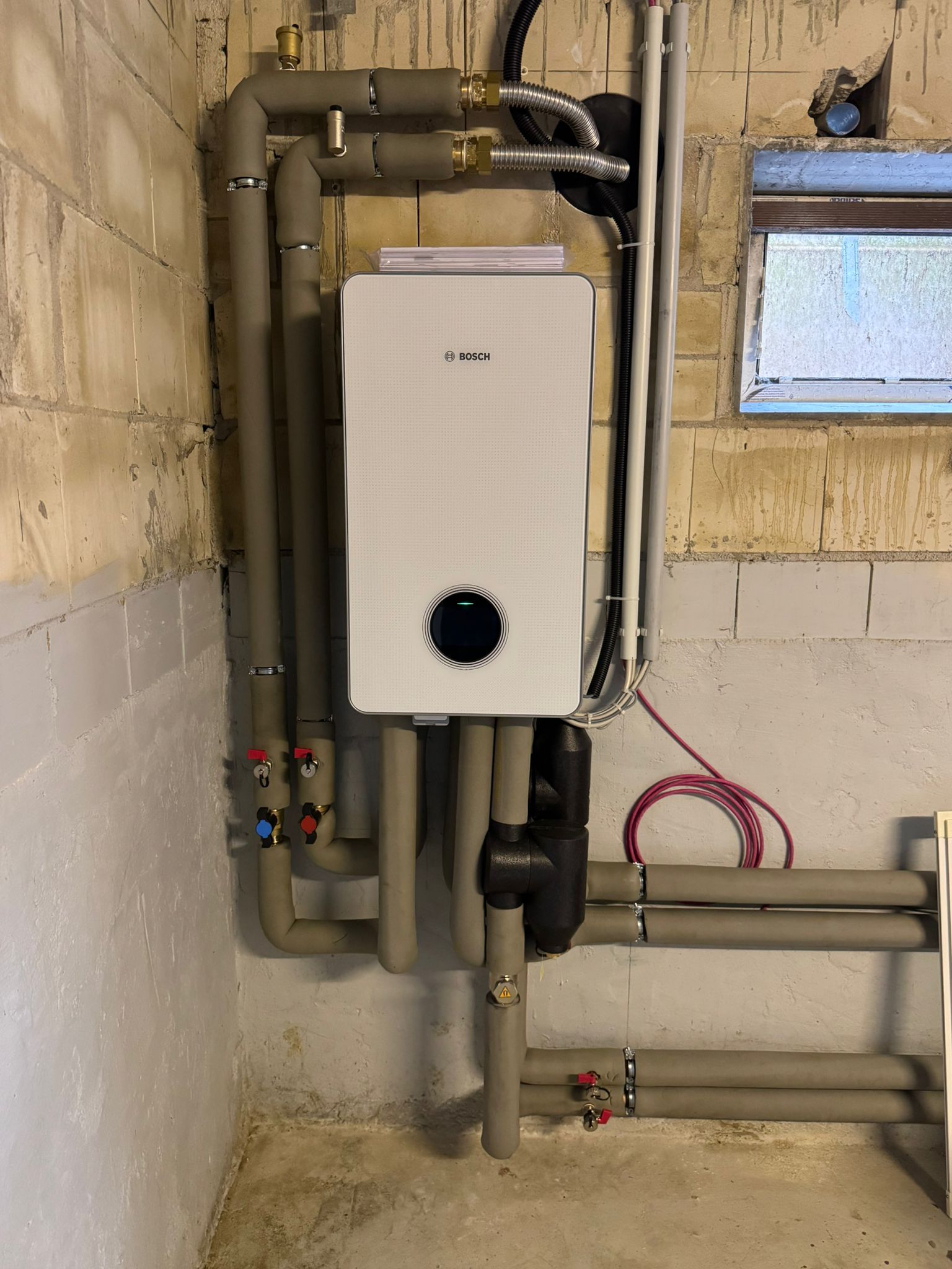 Bosch Inneneinheit im Keller, angeschlossen an isolierte Heizungsrohre und Steuerleitungen.
