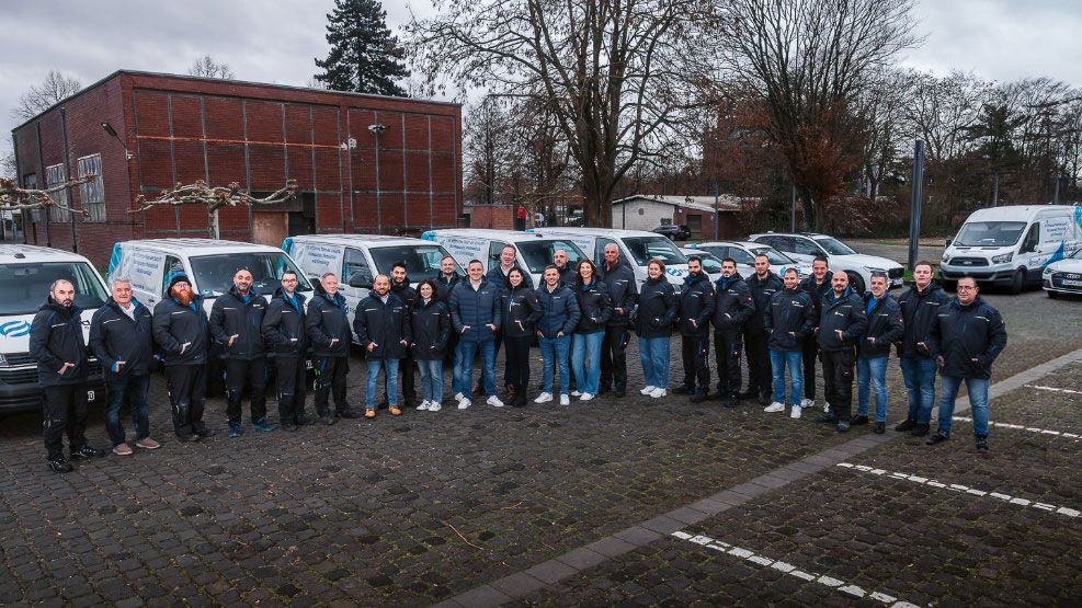 Neunköpfiges Montageteam in Arbeitskleidung posiert selbstbewusst vor Einsatzfahrzeugen auf einem Industriegelände.