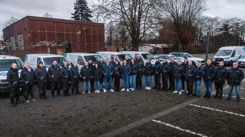 Team Friondo steht vor Firmenfahrzeugen auf einem Parkplatz, Gruppenfoto vor Backsteingebäude im Herbst.
