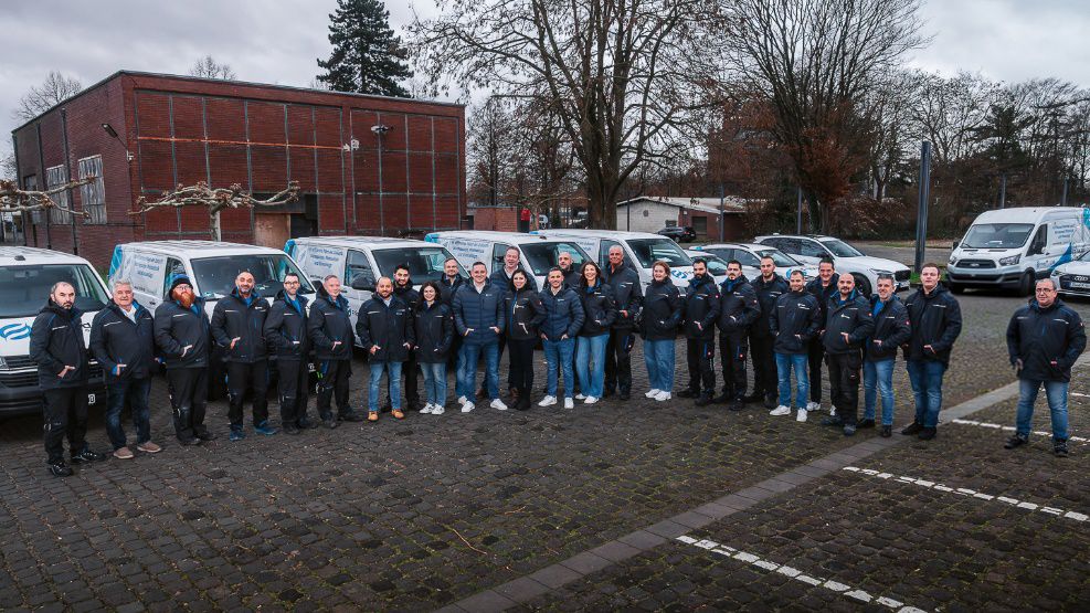 Neunköpfiges Montageteam in Arbeitskleidung posiert selbstbewusst vor Einsatzfahrzeugen auf einem Industriegelände.