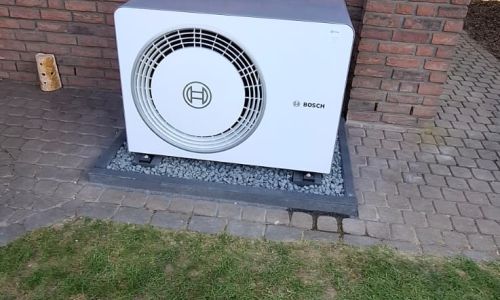 Bosch-Außengerät auf Terrasse mit grauen Fliesen, vor roter Ziegelwand neben Glastür und Blumenbeet.