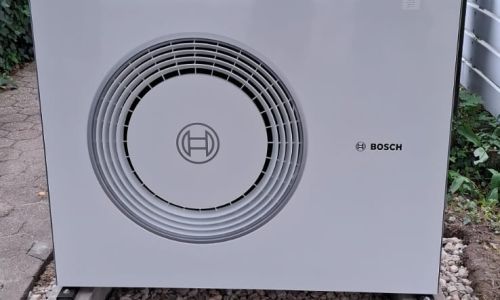 Außeneinheit einer Bosch-Wärmepumpe vor einem Wohnhaus, installiert auf Kiesbett mit Betonfundamenten im Vorgarten.