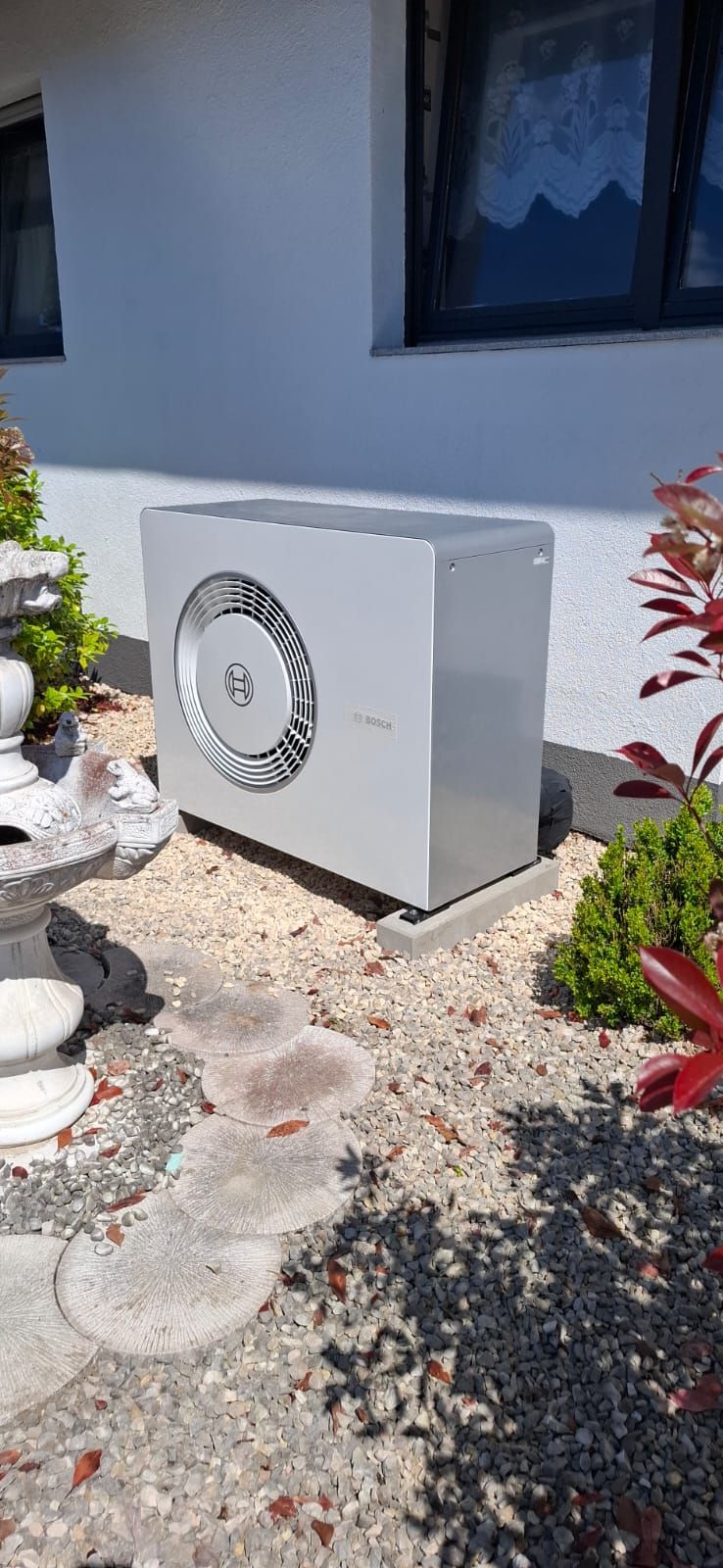 Außeneinheit einer Bosch-Wärmepumpe an weißer Hauswand, eingebettet in Kiesfläche mit Dekosteinen und Zierbrunnen.