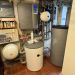 Technikraum mit Pufferspeicher, Wandgerät, zwei Ausdehnungsgefäßen und zahlreichen Rohrverbindungen.