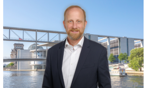 Ein Mann im Anzug steht vor dem Berliner Regierungsviertel mit Spree, Reichstag und moderner Brücke im Hintergrund.