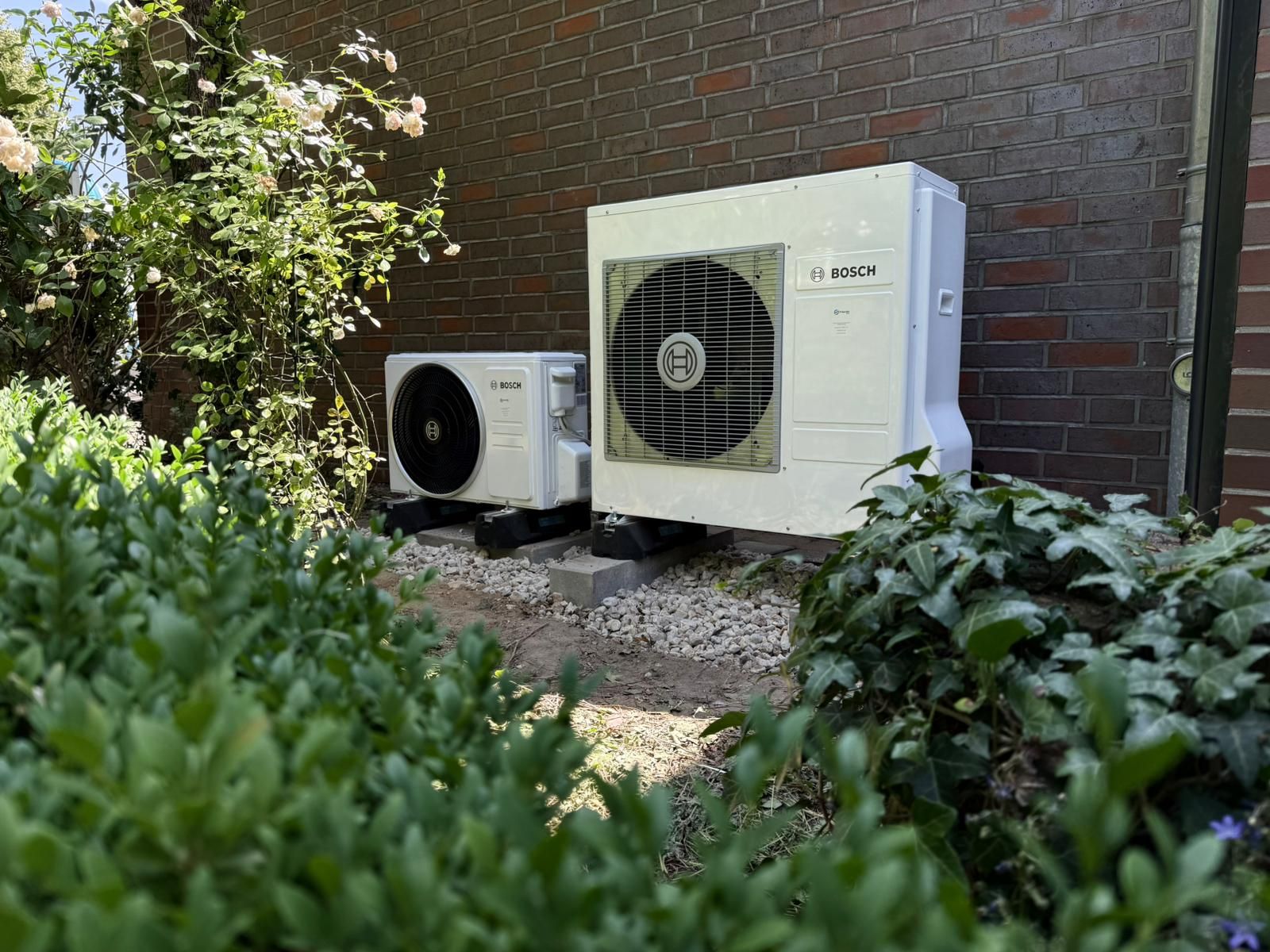 Bosch Außeneinheiten der Klimaanlage im Gartenbereich, umgeben von Sträuchern vor Hauswand.