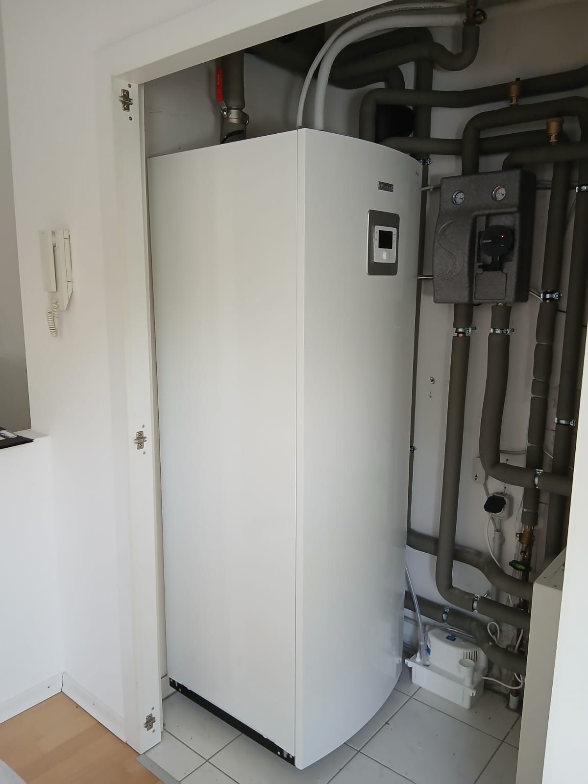Weiße Wärmepumpen-Inneneinheit mit Anschlüssen und Rohren in einem schmalen Technikschrank.