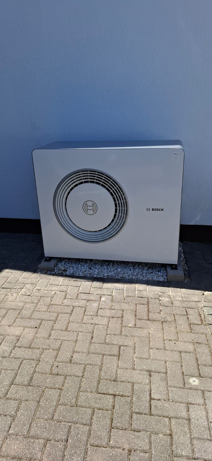 Außenstehende Bosch-Wärmepumpe an Hauswand auf Kiesbett mit Pflastersteinen im Vordergrund bei Tageslicht.