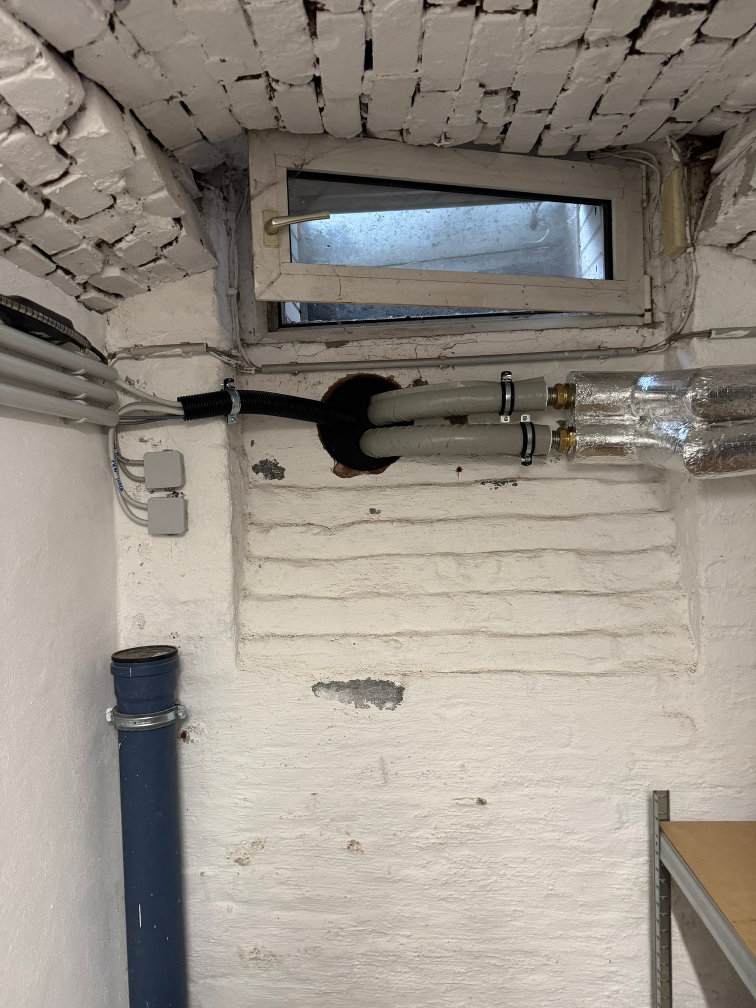 Kellerfenster mit durchgeführten Leitungen und gedämmten Rohren an weißer Wand.