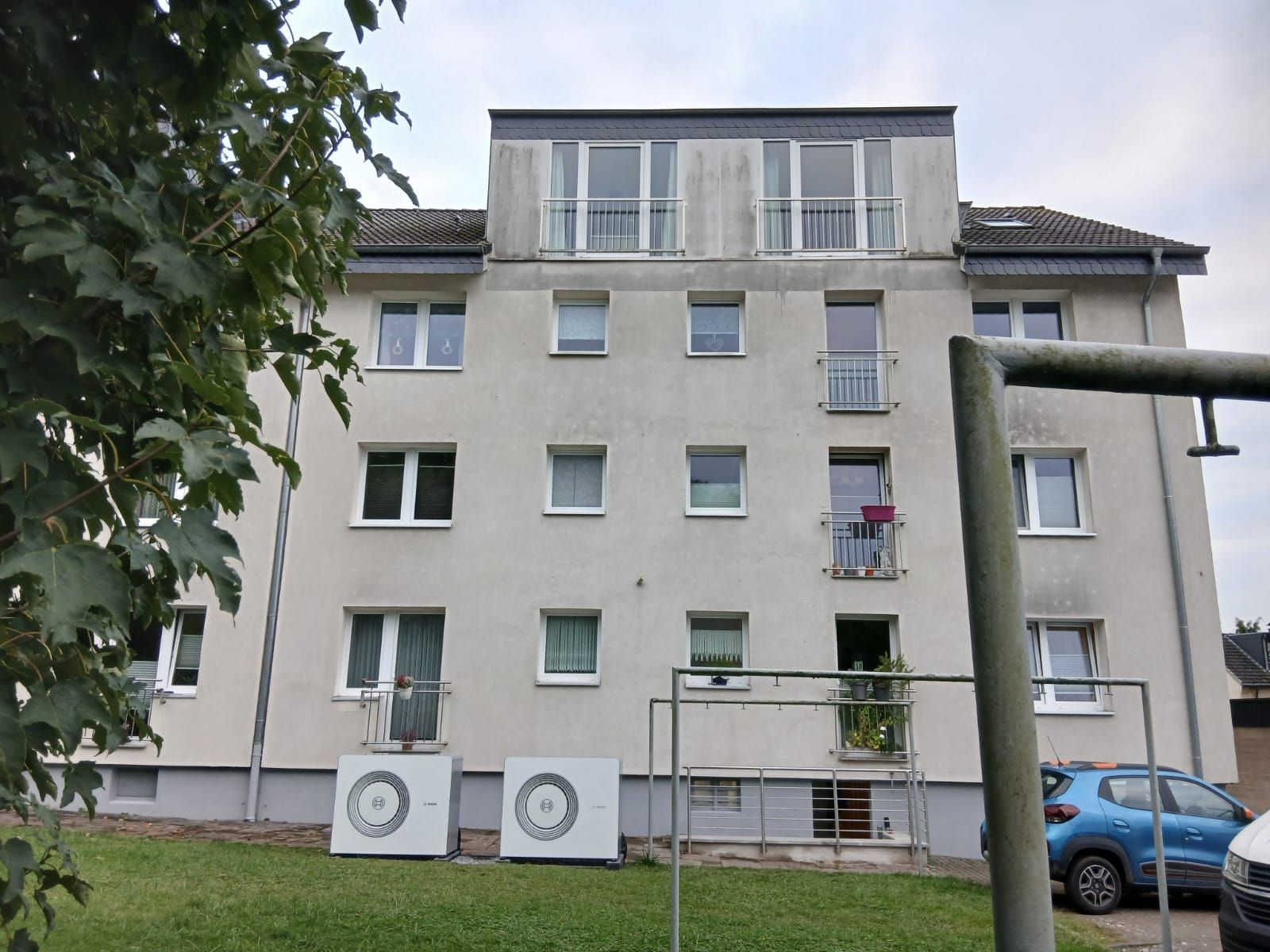 Außenansicht Mehrfamilienhaus, zwei Wärmepumpen auf Fundamenten im Gartenbereich.