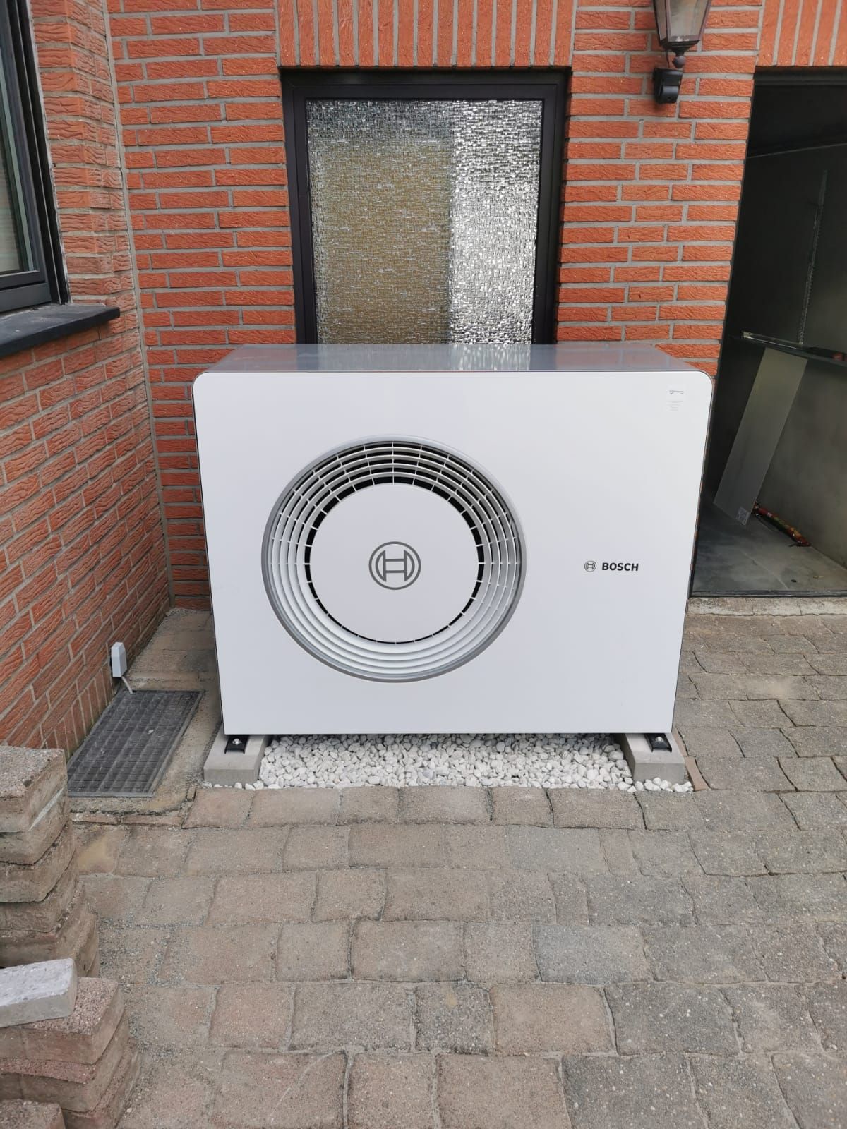 Bosch-Wärmepumpe vor roter Ziegelfassade auf gepflastertem Hof mit Kiesbett und Glasfenster im Hintergrund.