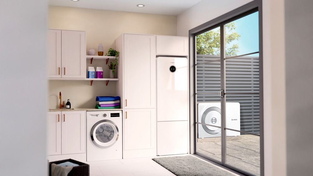 Moderner Hauswirtschaftsraum mit Waschmaschine, weißen Einbauschränken und sichtbarer Wärmepumpe im Außenbereich durch das Fenster.