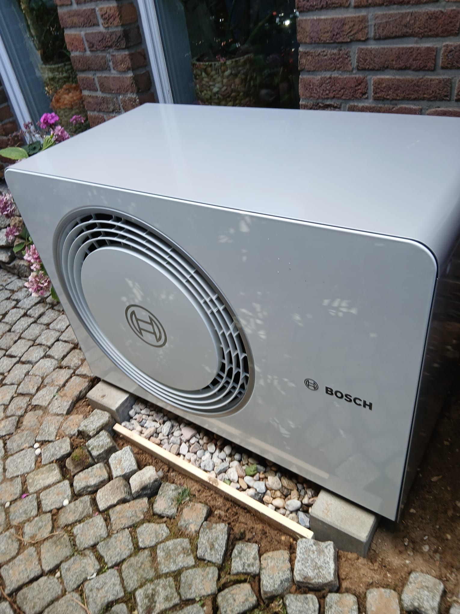 Außeneinheit einer Bosch-Wärmepumpe vor einem Wohnhaus, installiert auf Kiesbett mit Kiesfundament im Vorgarten.