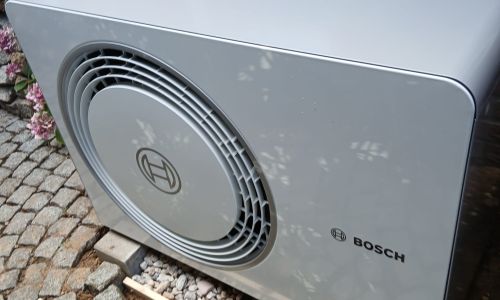 Außeneinheit einer Bosch-Wärmepumpe vor einem Wohnhaus, installiert auf Kiesbett mit Kiesfundament im Vorgarten.