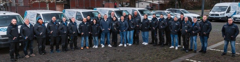 Neunköpfiges Montageteam in Arbeitskleidung posiert selbstbewusst vor Einsatzfahrzeugen auf einem Industriegelände.