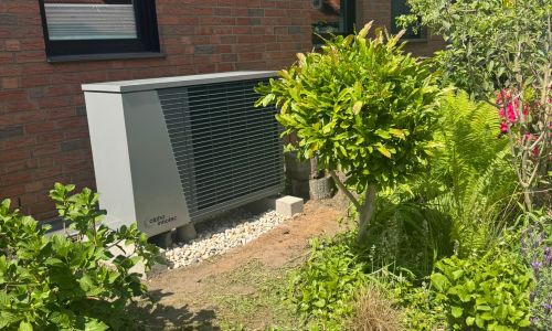 Außeneinheit einer Wärmepumpe vor roter Klinkerfassade, aufgestellt im bepflanzten Garten.