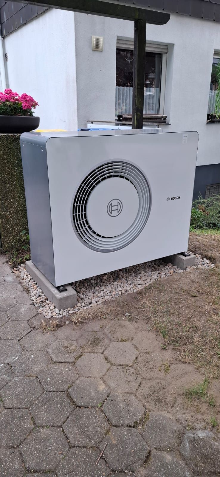 Außeneinheit einer Bosch-Wärmepumpe vor einem Wohnhaus, installiert auf Kiesbett mit Betonfundamenten im Vorgarten.