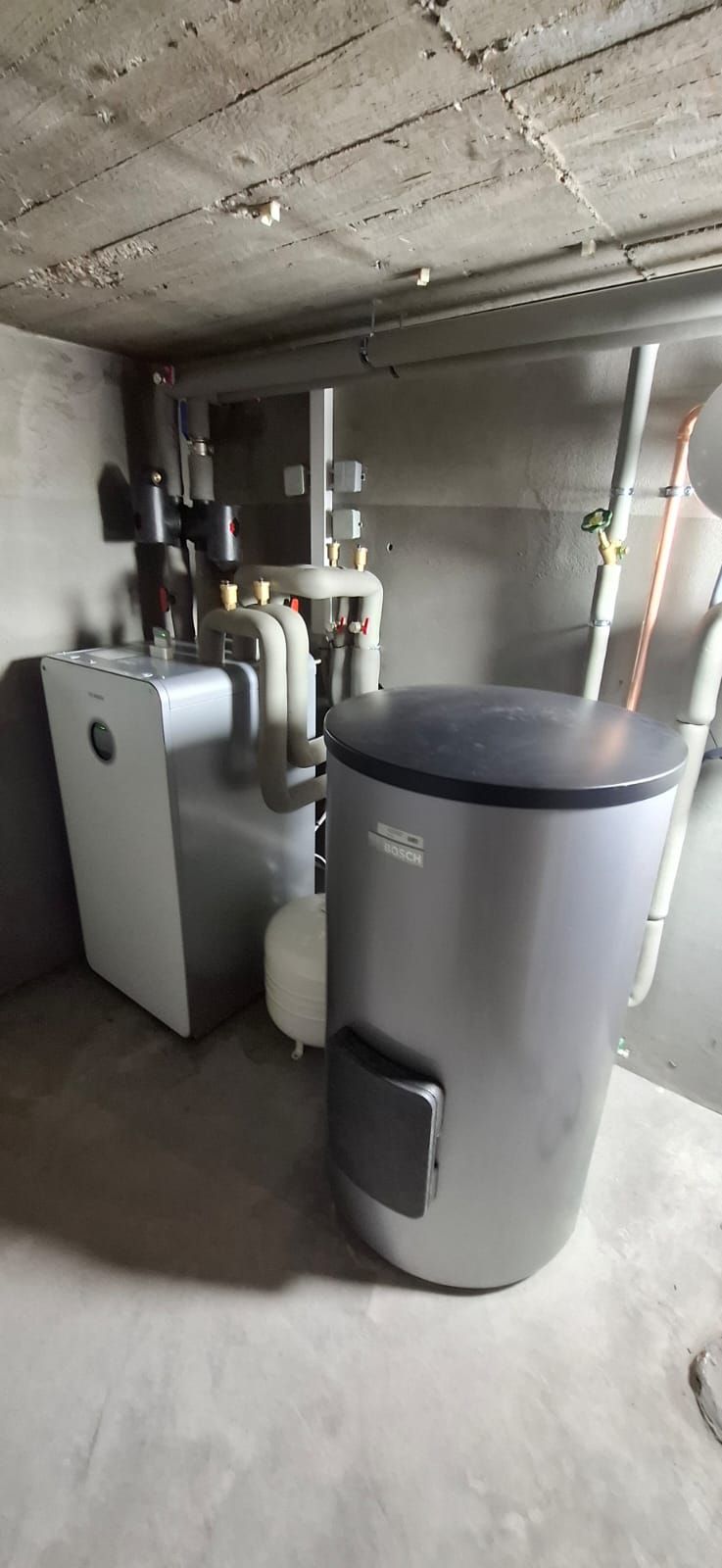 Wärmepumpen-Inneneinheit mit Pufferspeicher in grauem Technikraum, isolierte Rohre verlaufen an Wand und Decke.