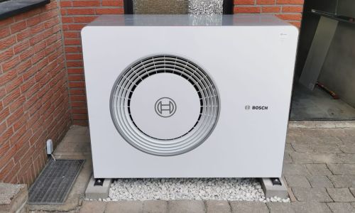 Bosch-Wärmepumpe vor roter Ziegelfassade auf gepflastertem Hof mit Kiesbett und Glasfenster im Hintergrund.
