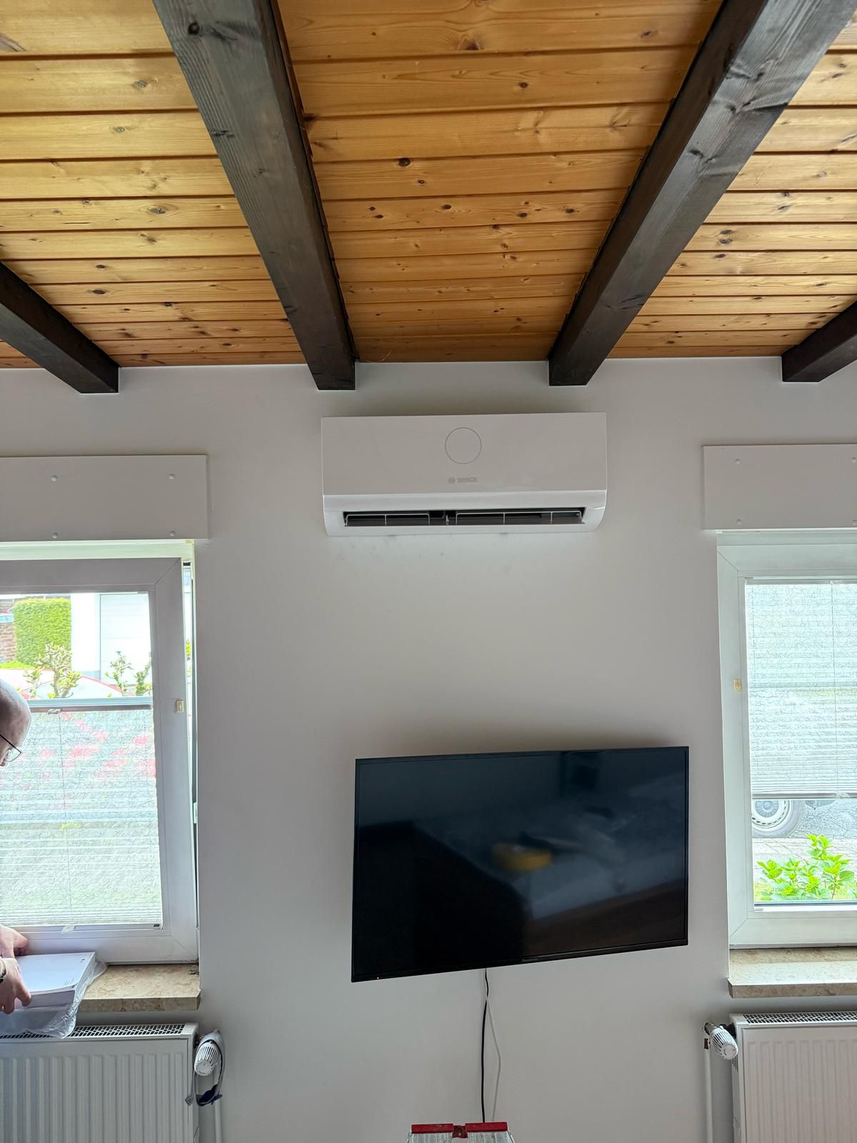 Wandmontiertes Bosch Klimagerät über Fernseher, sichtbar zwischen zwei Fenstern unter Holzbalkendecke.