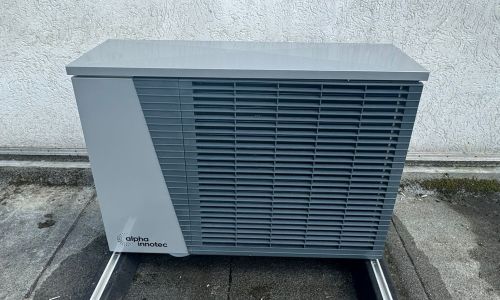 Bosch-Wärmepumpe außen auf Kiesbett vor weißer Hauswand, neben Gehweg und Sträuchern, mit Werkzeug im Vordergrund.