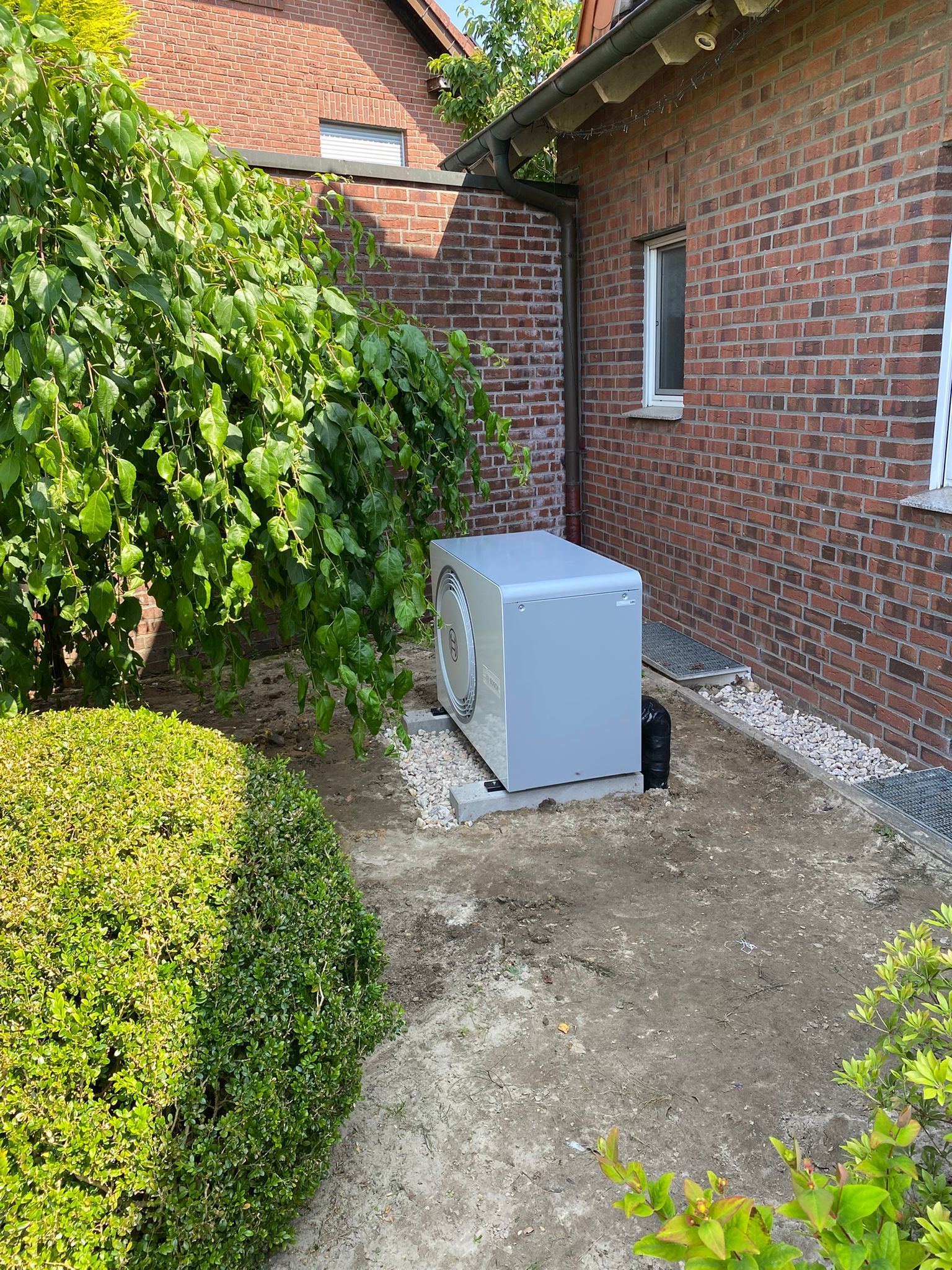 Außengerät einer Wärmepumpe auf Kiesbett vor rotem Ziegelhaus, von Büschen und Bäumen umgeben.