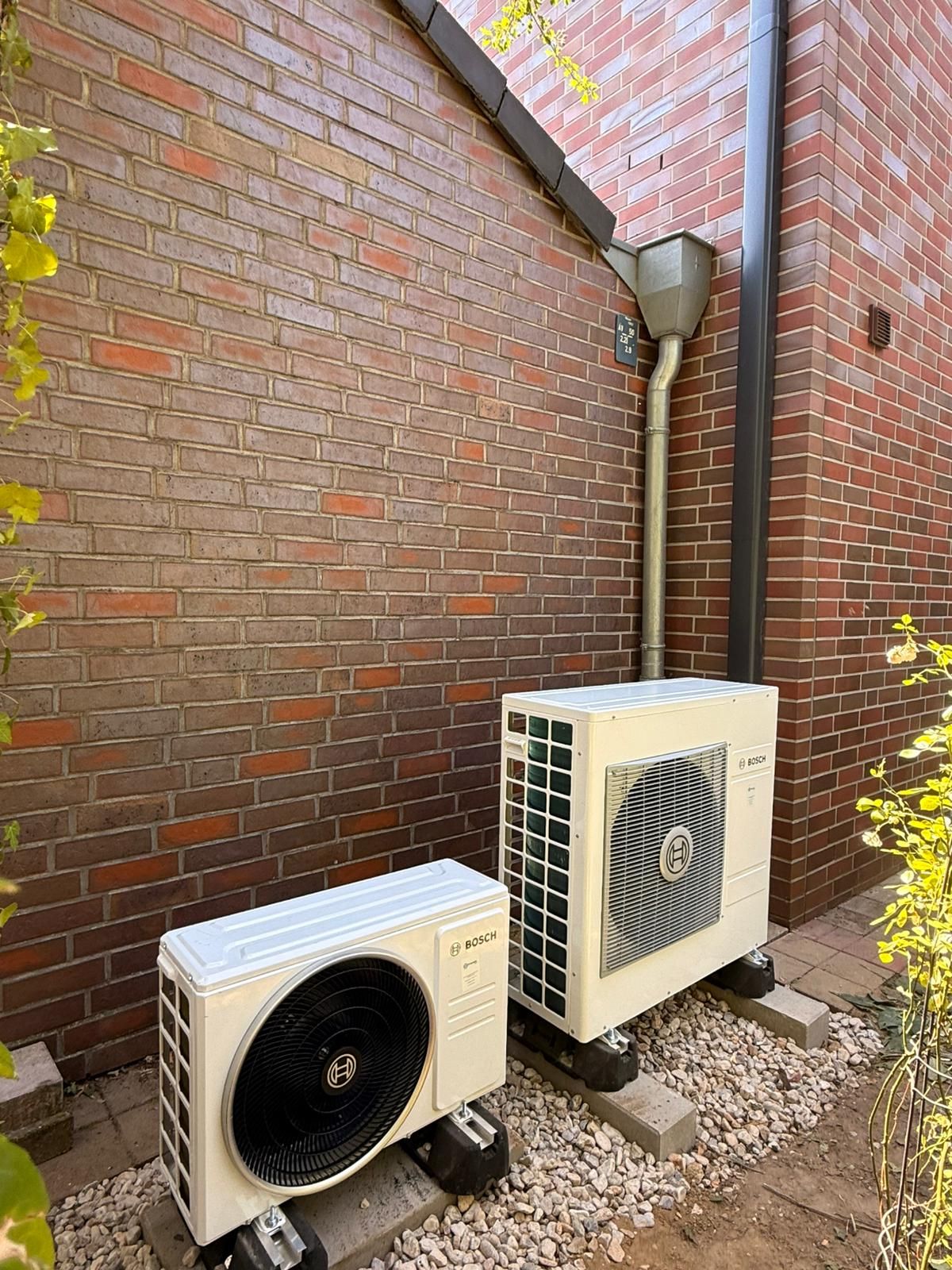 Zwei Bosch Klima Außengeräte auf Betonfüßen vor roter Klinkerfassade neben Fallrohr.