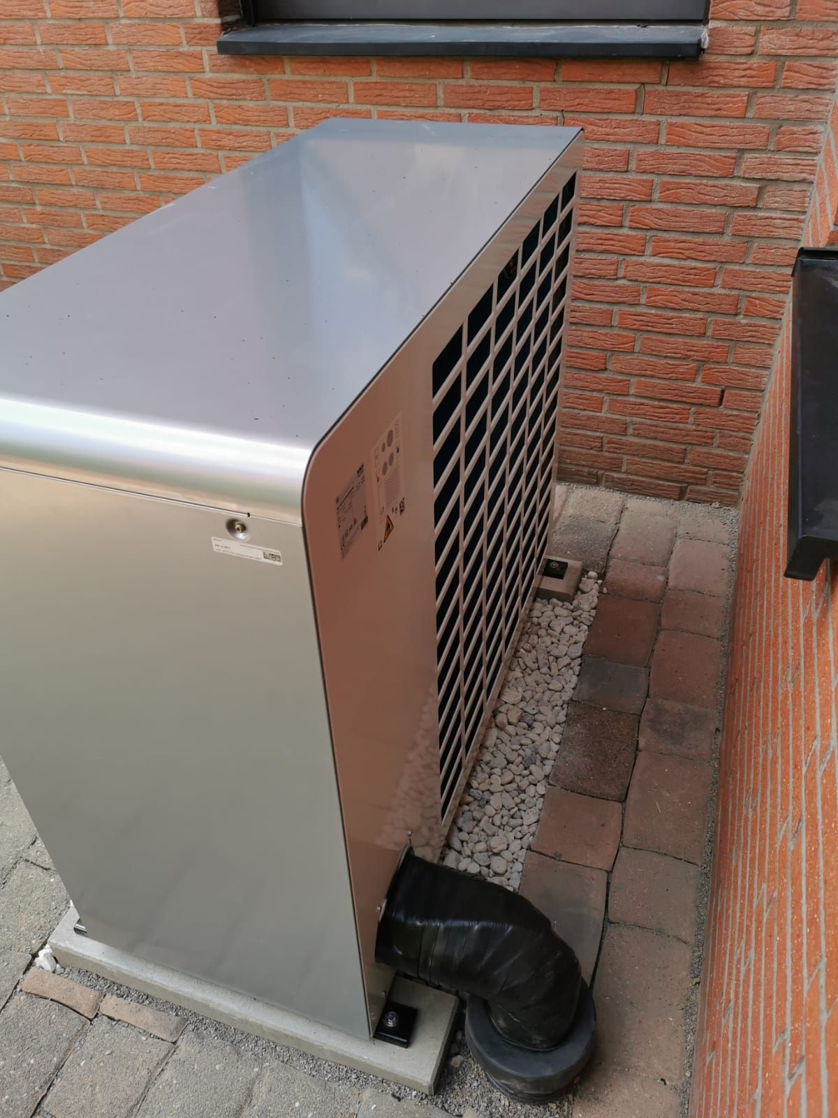Rückseite einer Wärmepumpe mit Lüftungsgitter und Isolierschlauch, auf Kiesbett zwischen zwei Ziegelwänden.