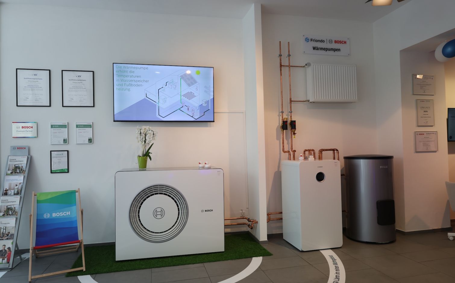 Moderne Wärmepumpe von Bosch an einer dunklen Hauswand im Garten, mit Blumenbeeten und Holzschuppen im Hintergrund.
