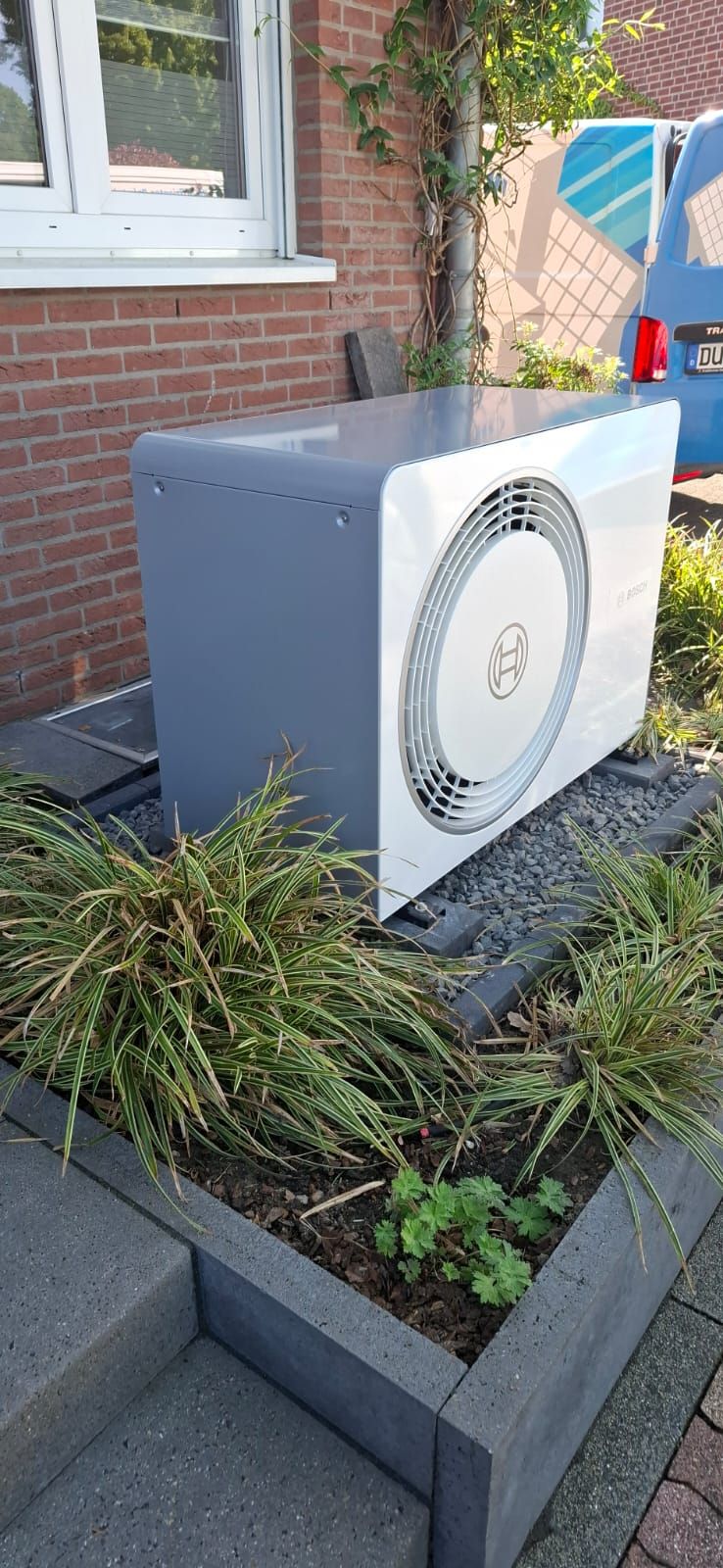 Außenstehende Bosch-Wärmepumpe auf Kiesbett vor roter Ziegelwand und Fenster, mit Pflanzen umgeben.