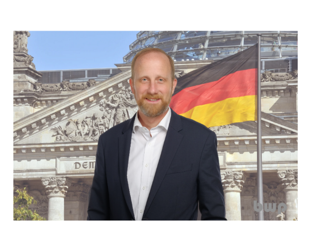 Ein Mann im Anzug steht vor dem Reichstagsgebäude in Berlin, daneben weht eine große Deutschlandfahne.