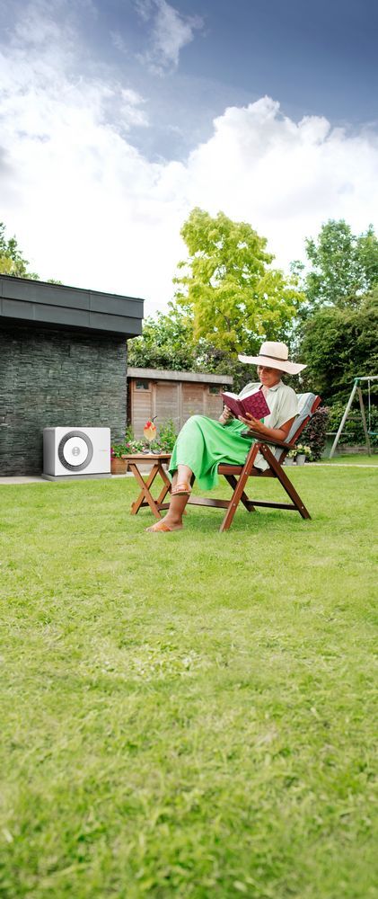 Frau liest entspannt im Garten, im Hintergrund steht eine weiße Bosch-Wärmepumpe an der dunklen Hausfassade.