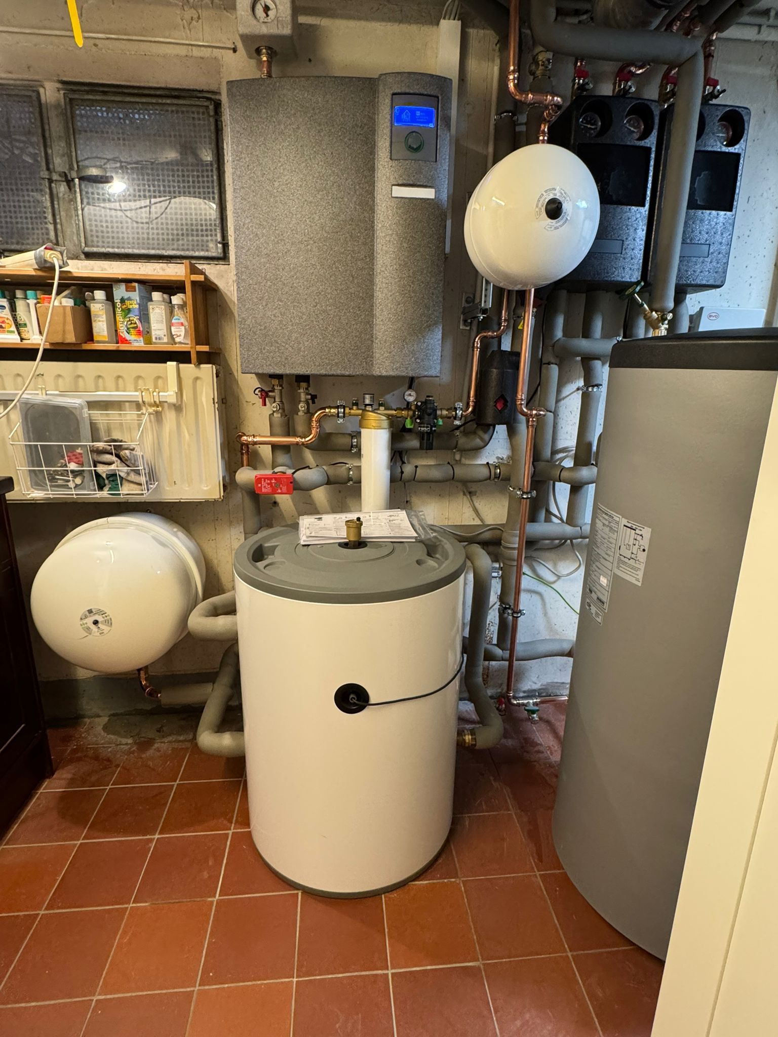 Technikraum mit Pufferspeicher, Wandgerät, zwei Ausdehnungsgefäßen und zahlreichen Rohrverbindungen.