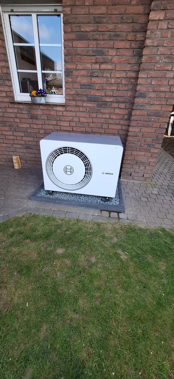 Bosch-Außengerät auf Terrasse mit grauen Fliesen, vor roter Ziegelwand neben Glastür und Blumenbeet.