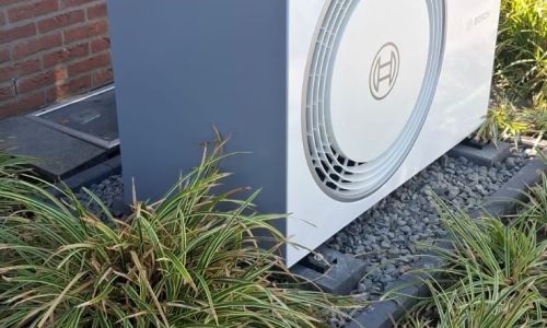 Außenstehende Bosch-Wärmepumpe auf Kiesbett vor roter Ziegelwand und Fenster, mit Pflanzen umgeben.