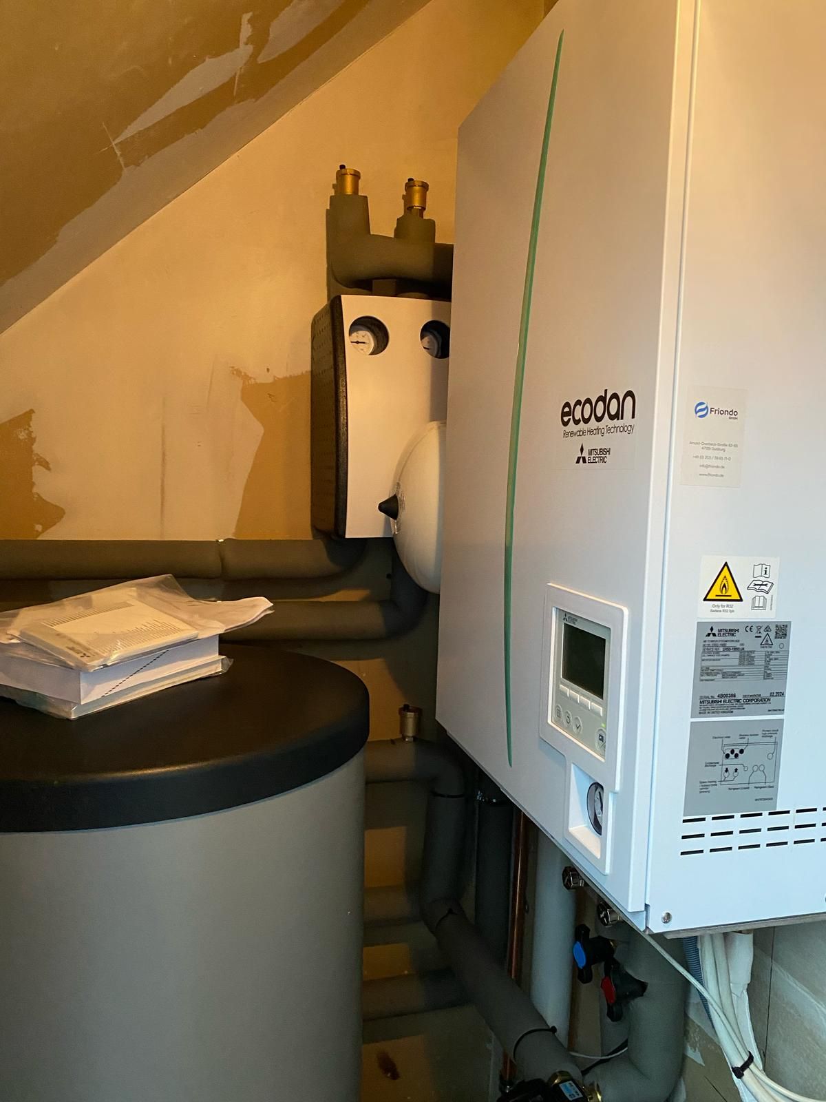 Ecodan-Wärmepumpenmodul an Wand mit Pufferspeicher, Isolierungen und aufliegenden Bedienungsanleitungen.
