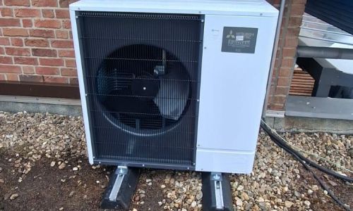 Außengerät einer Mitsubishi Zubadan-Wärmepumpe auf Schotterfläche vor roter Klinkerwand mit Dachschräge.