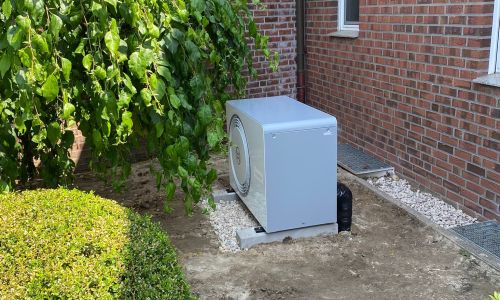 Außengerät einer Wärmepumpe auf Kiesbett vor rotem Ziegelhaus, von Büschen und Bäumen umgeben.