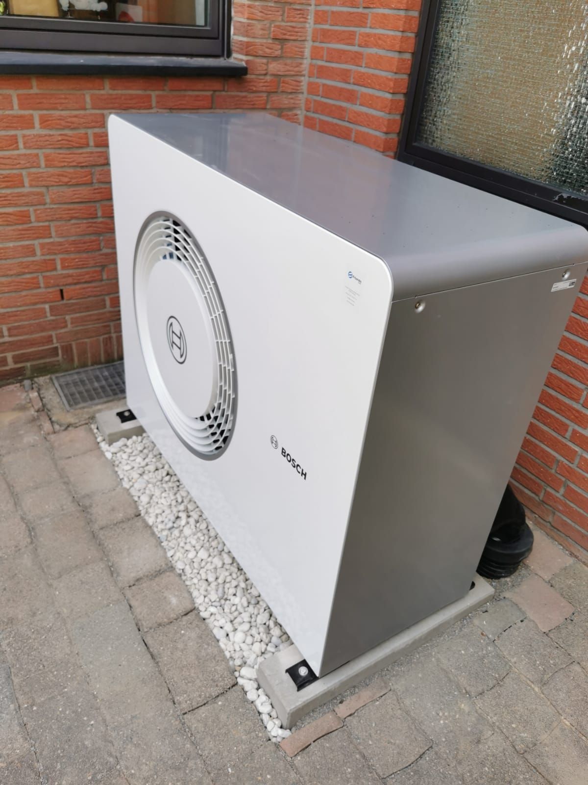 Bosch-Wärmepumpe vor roter Ziegelfassade auf gepflastertem Hof mit Kiesbett und Glasfenster im Hintergrund.