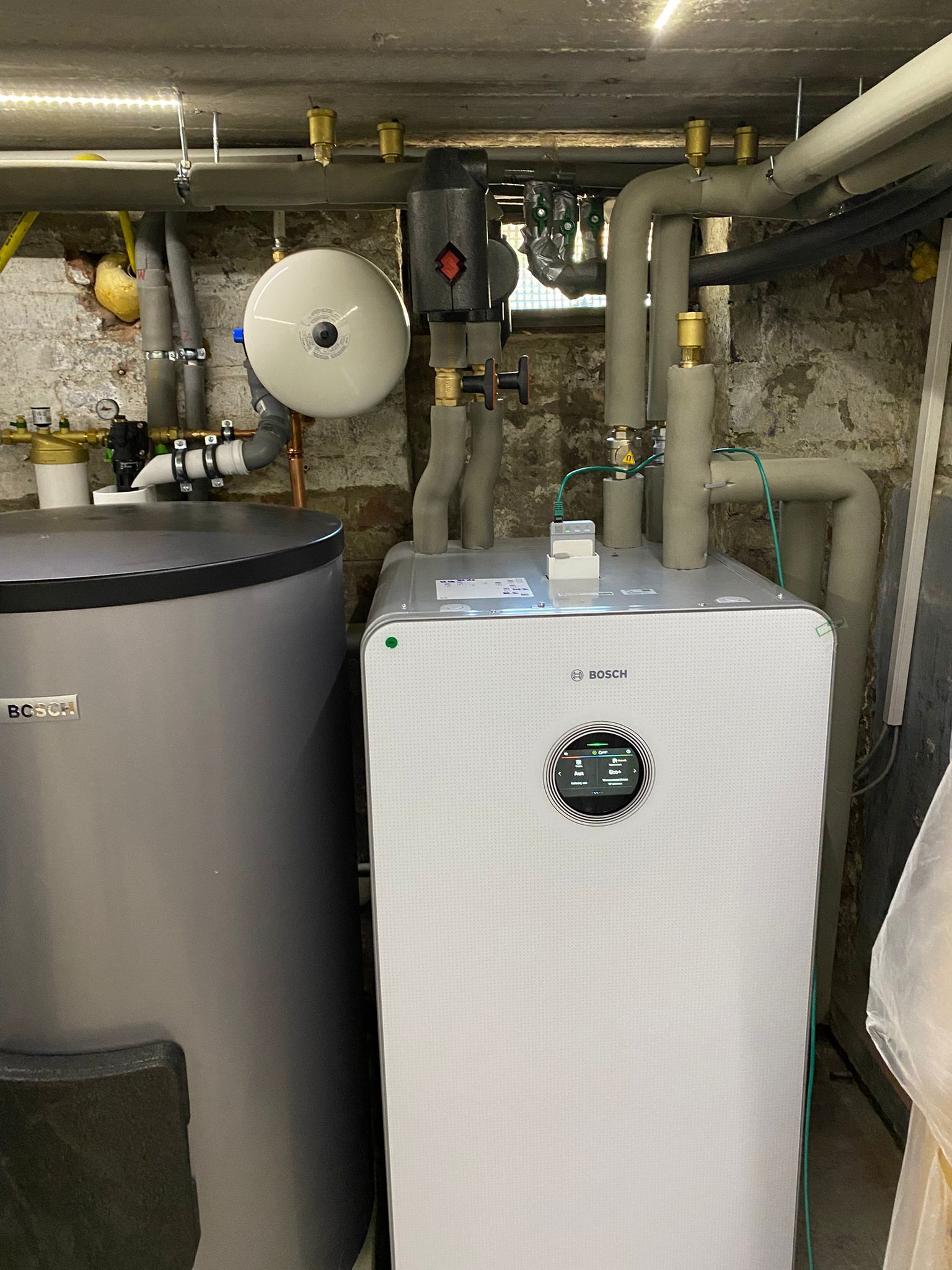 Bosch Wärmepumpen Inneneinheit im Keller neben Pufferspeicher, mit gedämmten Rohrleitungen und Anzeige.