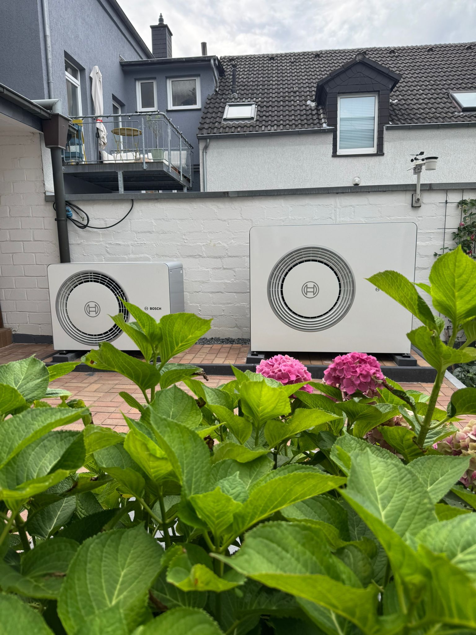 Zwei Bosch Wärmepumpen Außengeräte vor weißer Gartenmauer, umgeben von Pflanzen.