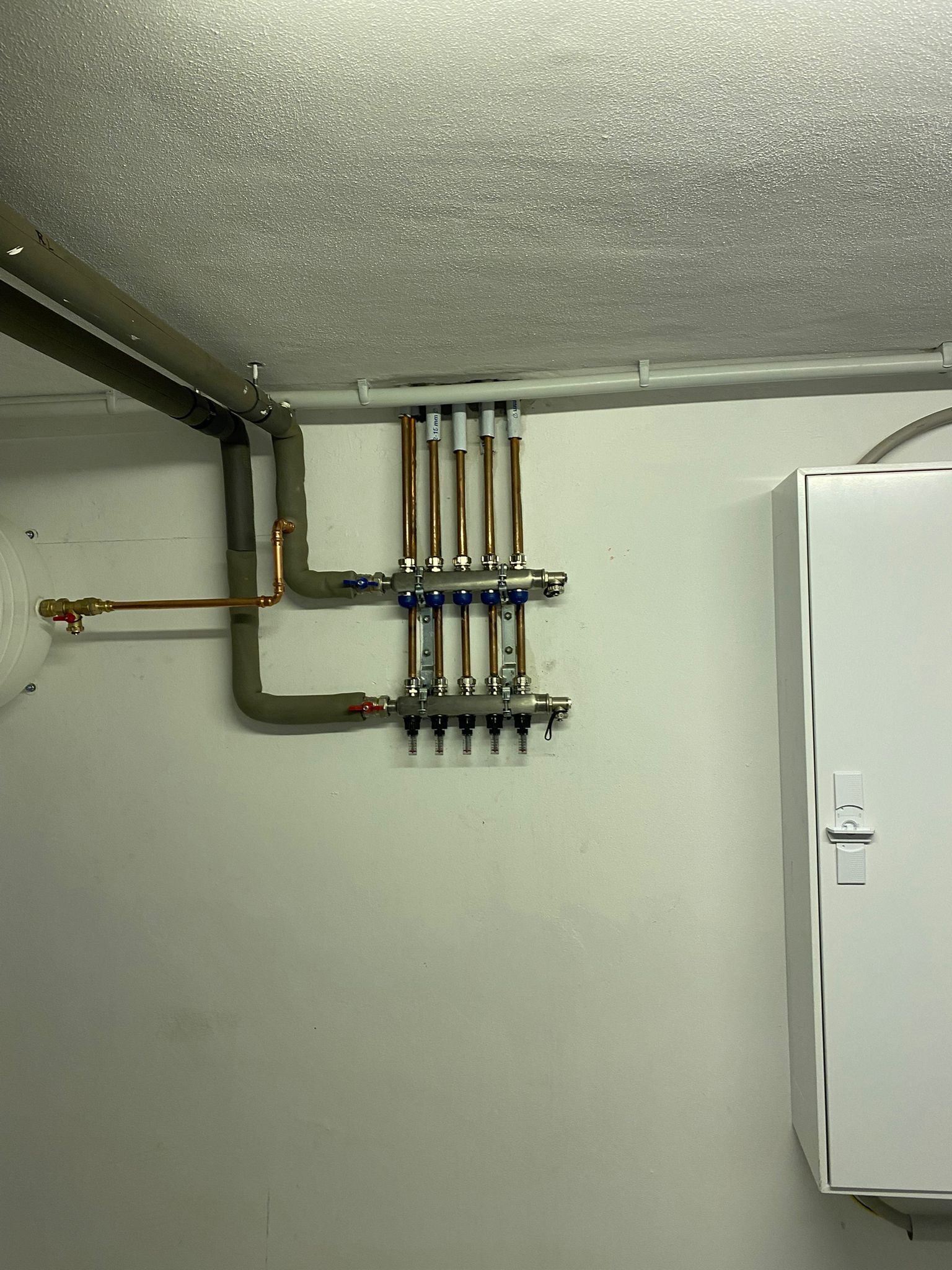 Rohrverteiler mit sechs Kupferrohren an weißer Wand, angeschlossen an isolierte Vor- und Rücklaufleitungen.