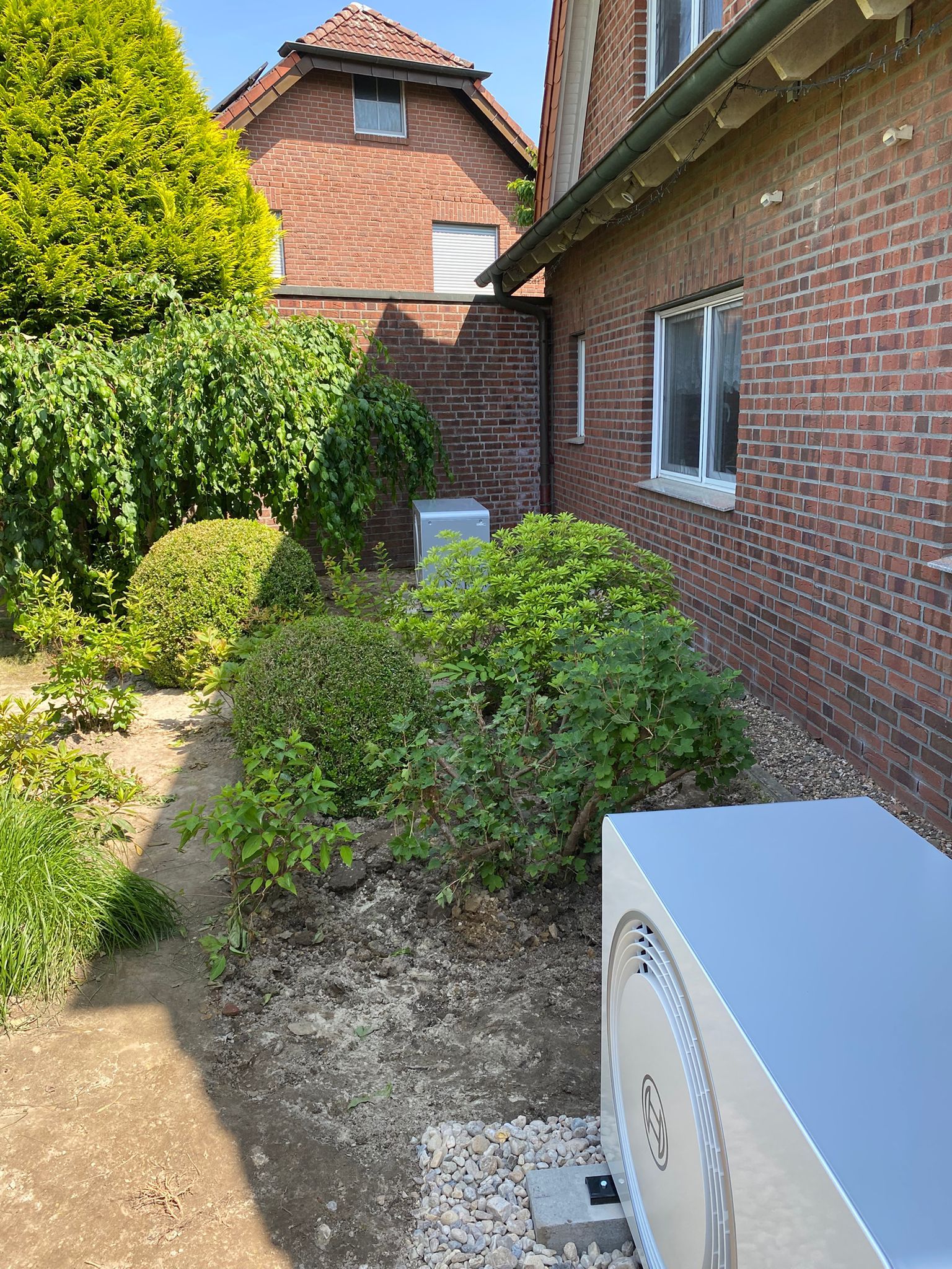 Zwei Außengeräte von Wärmepumpen auf Kiesbett vor rotem Ziegelhaus, von Büschen und Bäumen umgeben.