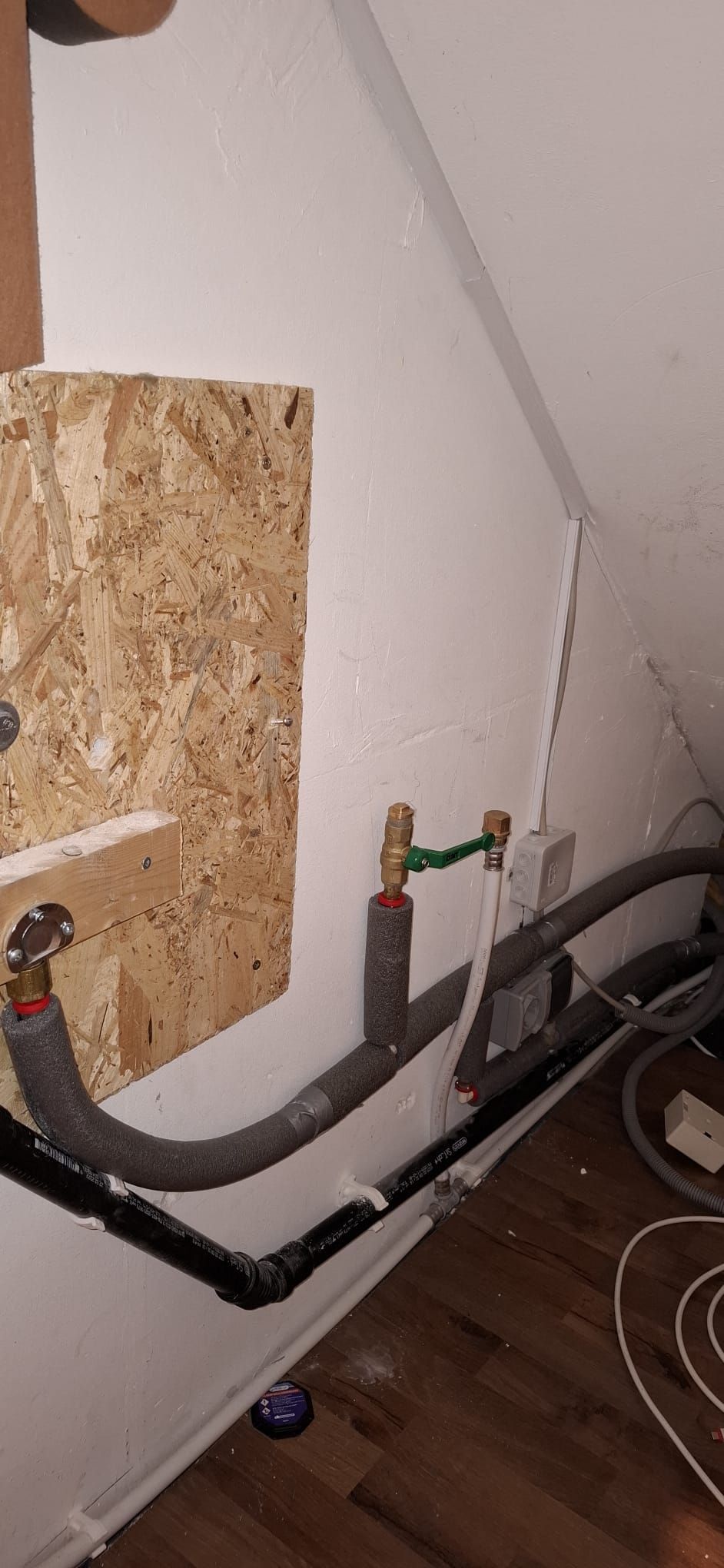Wasseranschlüsse mit Isolierung und Absperrhähnen unter Treppe an weißer Wand mit Holzverkleidung.