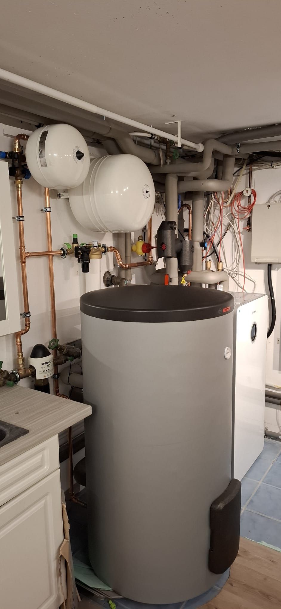 Bosch-Pufferspeicher mit Heizungseinheit, Rohren und Wandarmaturen in Technikraum mit gefliestem Boden.