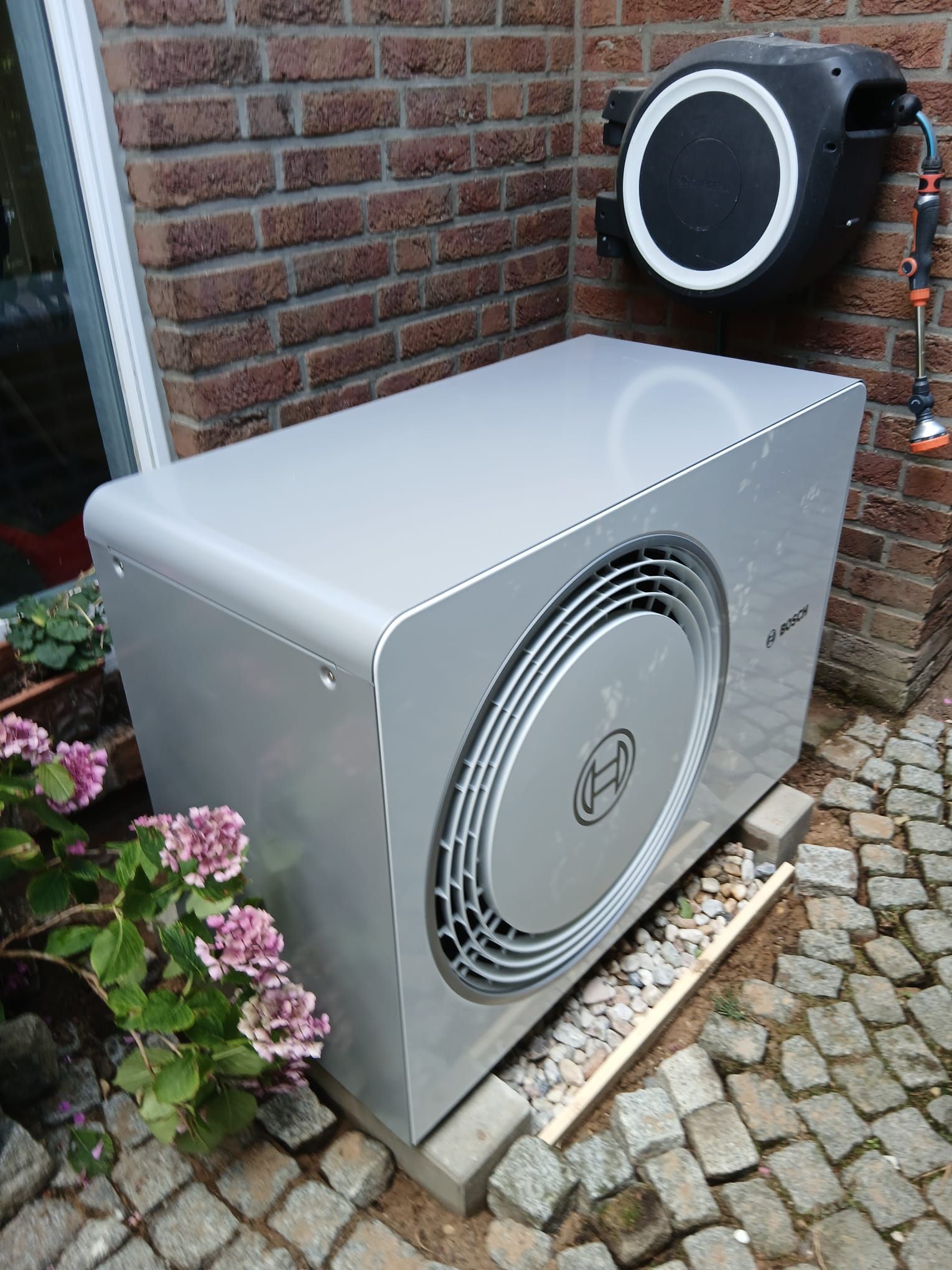 Außeneinheit einer Bosch-Wärmepumpe vor einem Wohnhaus, installiert auf Kiesbett mit Kiesfundament im Vorgarten.