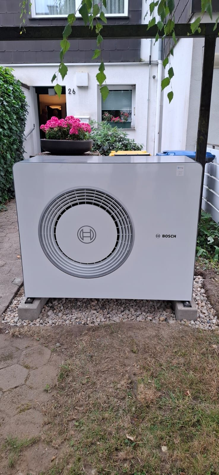 Außeneinheit einer Bosch-Wärmepumpe vor einem Wohnhaus, installiert auf Kiesbett mit Betonfundamenten im Vorgarten.
