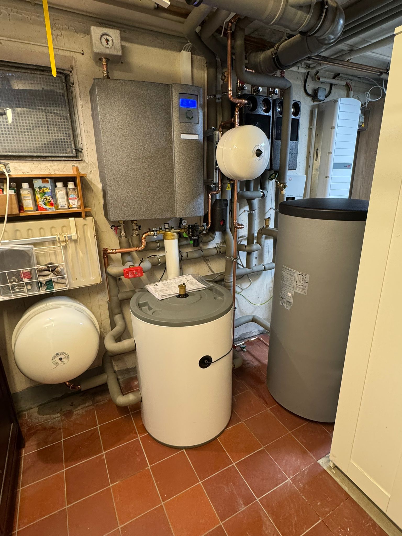 Technikraum mit Pufferspeicher, Wandgerät, zwei Ausdehnungsgefäßen und zahlreichen Rohrverbindungen.