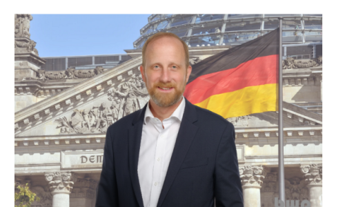 Ein Mann im Anzug steht vor dem Reichstagsgebäude in Berlin, daneben weht eine große Deutschlandfahne.