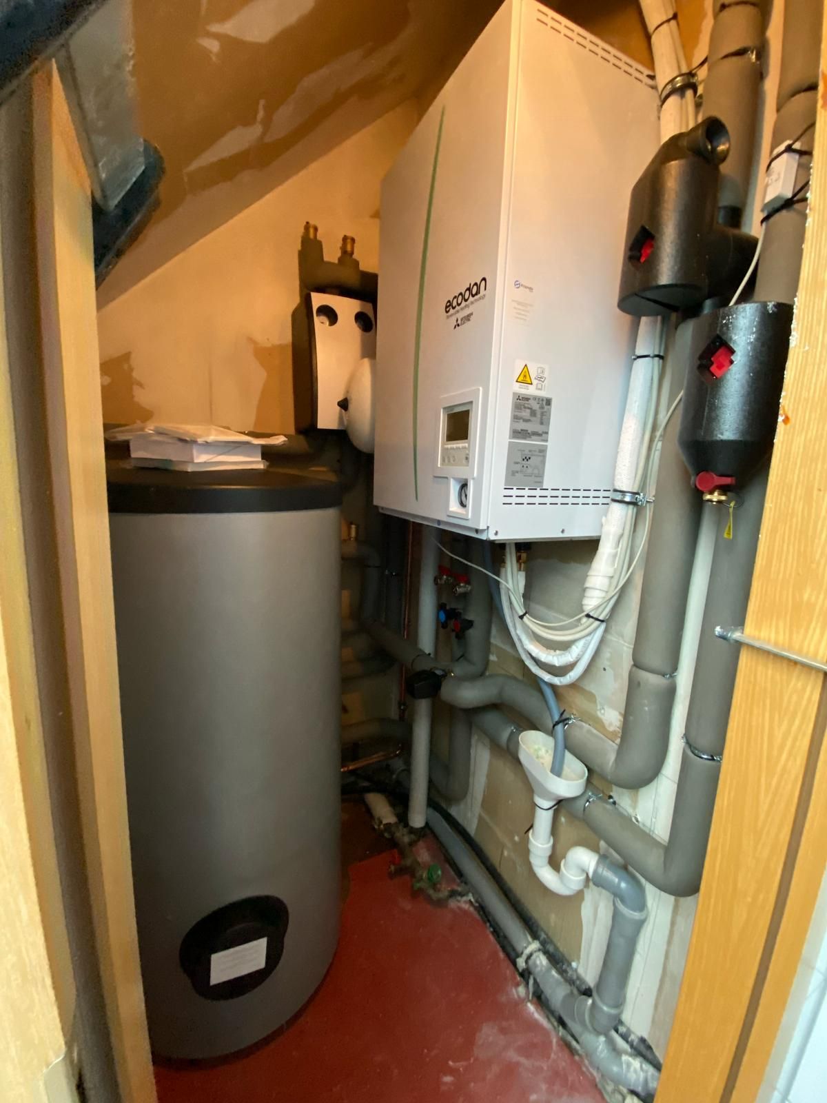 Ecodan-Wärmepumpenmodul an Wand mit Pufferspeicher, Isolierungen und aufliegenden Bedienungsanleitungen.